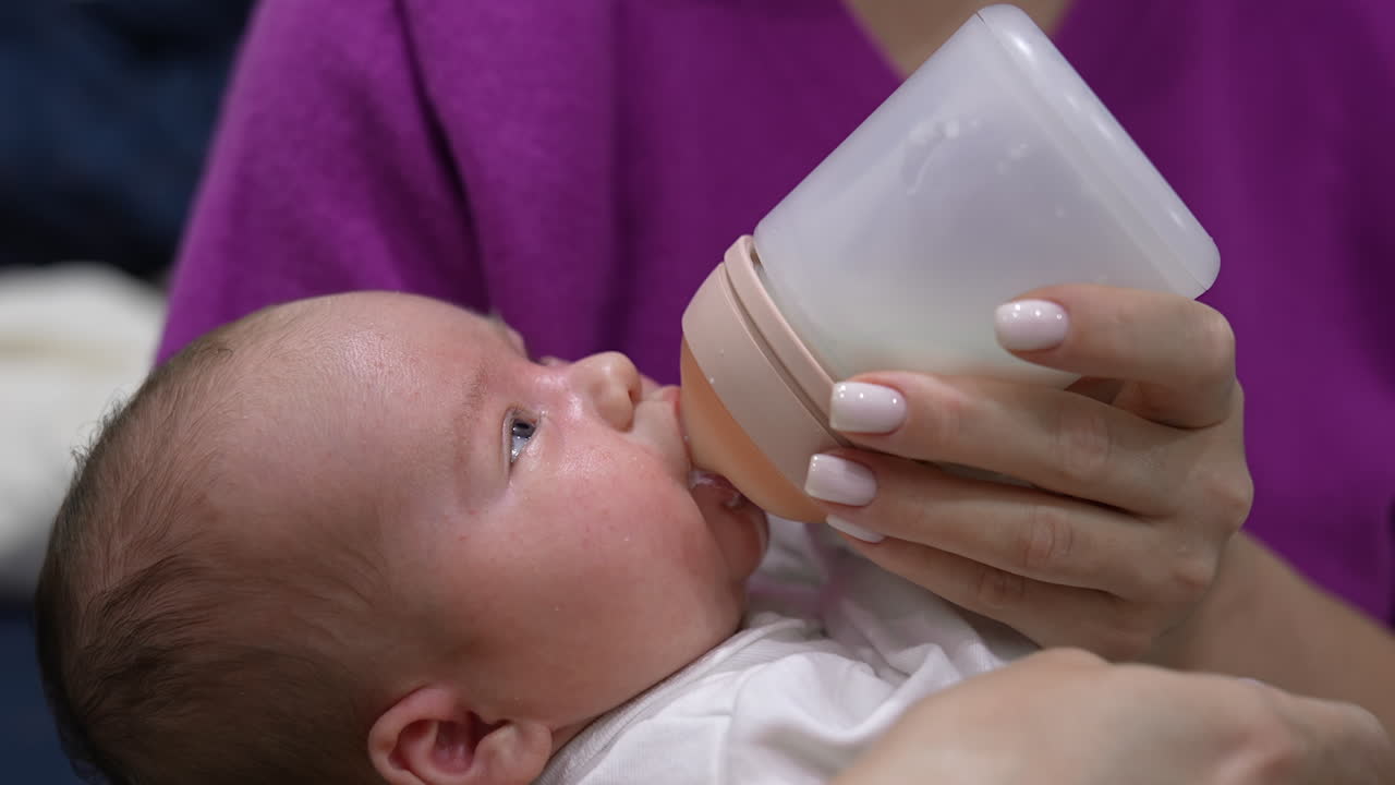 Little infant baby lying on mother's hands. Tiny baby suckling milk from the plastic bottle. Close up shot.