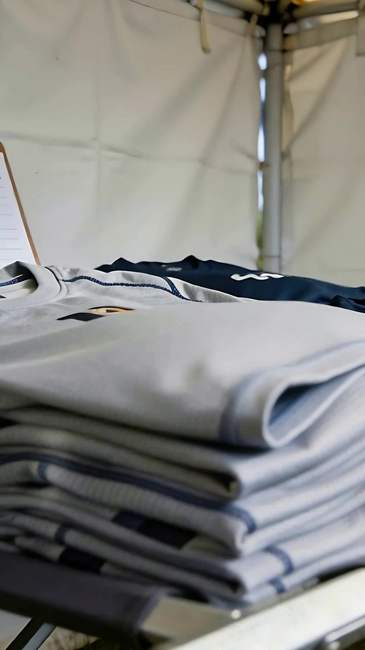 Video verticale: spostamento di una maglia su una pila grigia che si sistema su un tavolo pieghevole in una tenda, mostrando il logo dorato