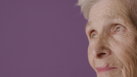 Close-Up-View-Of-Senior-Woman-Face-With-Short-Hair-And-Green-Eyes-Wearing-Earrings-Posing-At-Camera-On-Purple-Background