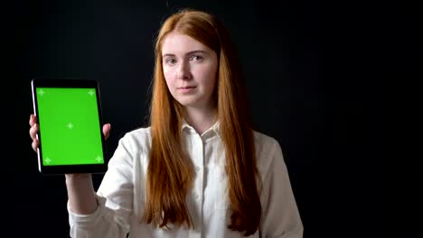 smiling ginger woman typing on tablet and showing screen with chromakey at camera, happy and cheerful, black background