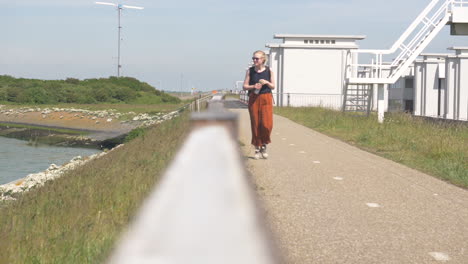 a young blonde female wearing sunglasses orange pants and blacktop walking along a footpath holding a bottle of water