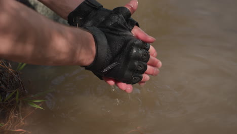 close-up view of tourist wearing black gloves while dipping and washing hands in muddy river water near grassy riverbank, sunlight casting warm glow on fingers and gentle ripples in water surface