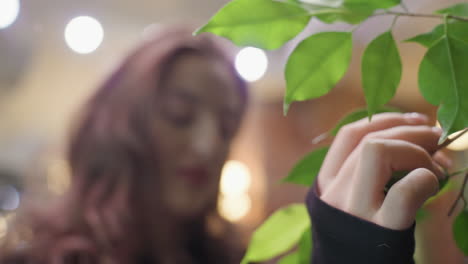 close up of female hand with acrylic nails gently holding vibrant green leaf while her face appears softly blurred in background, creating serene contrast between focus and softness in natural setting