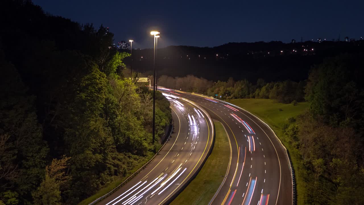 Zoom out long exposure time lapse of light trails headlights and tail lights from traffic on a curved highway road leading towards downtown Toronto at night with trees lining the sides of the road.