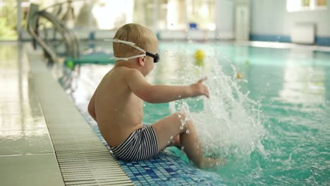 a small blond boy sits on the edge of the pool in swimming trunks and glasses and sprinkles water with his feet. splashing the water. having fun. indoors