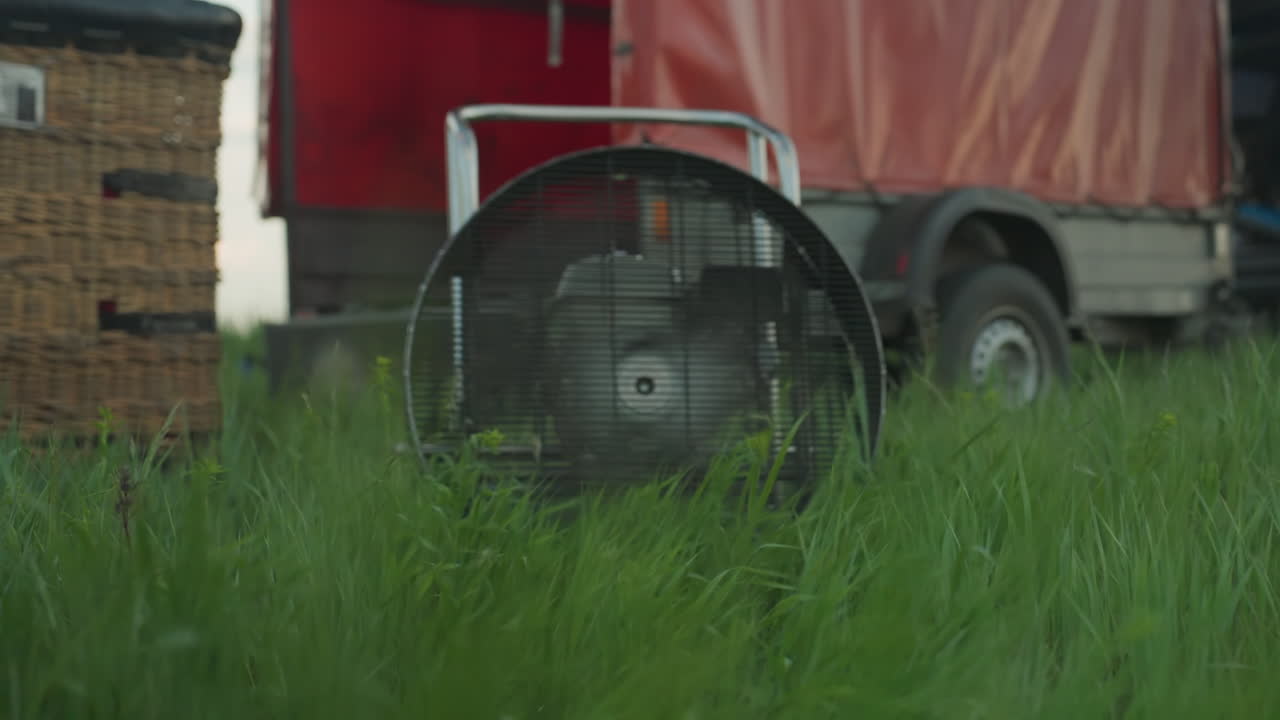man operating hot air balloon engine fan creating strong airflow that bends grass in foreground on grassy field with trailer and balloon envelope in background under cloudy sky