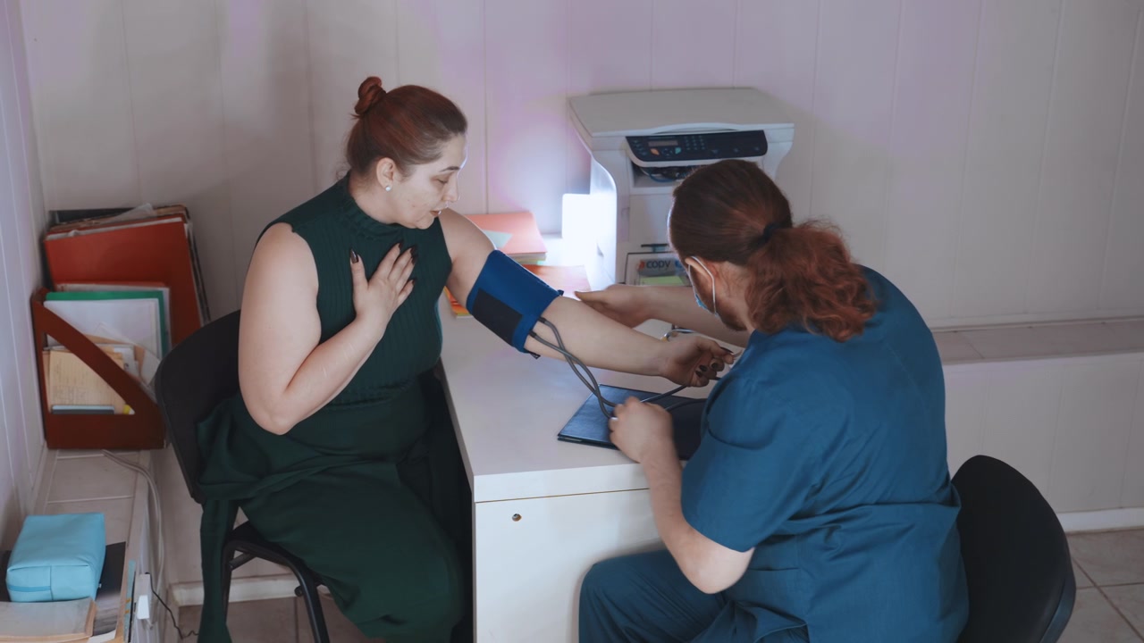 Hvid kvindelig patient vejledes gennem blodtryksmåling af hvid sygeplejerske, demonstration af manchetplacering, trin-for-trin instruktion, roligt hjemmekontormiljø med printer og formularer, patient.