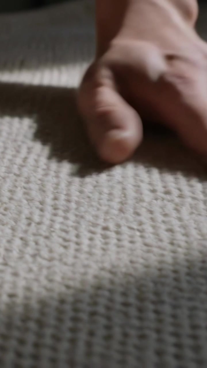Video verticale: Entra dall'alto a sinistra una sola mano che accarezza un plaid in maglia beige sul divano, lisciandolo.