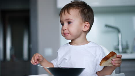 lovely child wearing white t-shirt sitting at table. baby boy holds a piece of bread and chews his food.