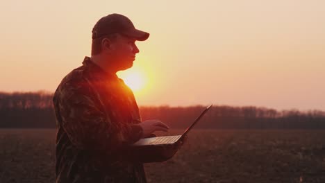 a middle-aged farmer uses a laptop in a field at sunset. plans sowing