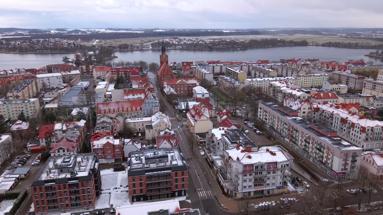 Snowfall in Elk city with colorful buildings roof covered in snow, Church of the sacred Heart of Jesus in view near Elk lake, Armii Krajowej street, Establishing drone shot, Panoramic