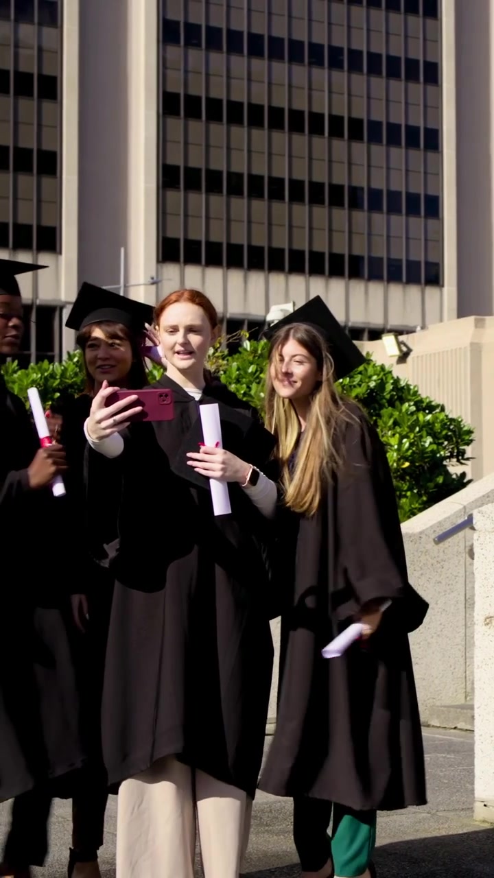 Vertical video: Descending grads in gowns on roof forming, redhead raising pink phone for selfie