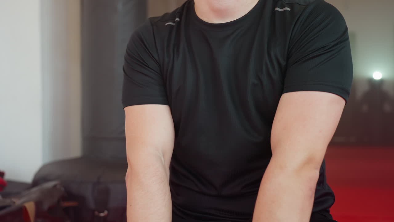 Boxer in black shirt adjusts black wrist wraps preparing for fight training, focusing on protection, discipline, and readiness for combat in gym environment with red mat and punching bags in background