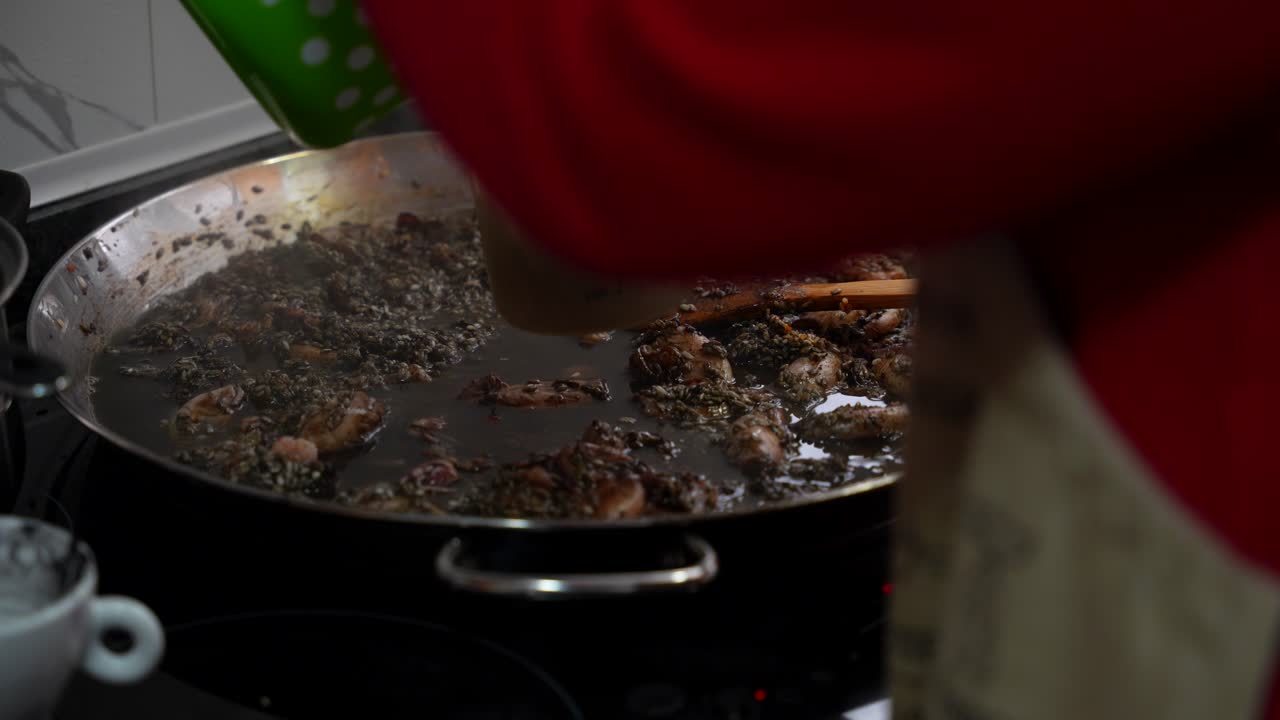 Woman pouring water on a paella to cook rice with black squid ink in the kitchen
