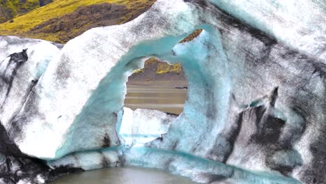 Während-Er-Sich-Mit-Einer-Drohne-In-Ein-Gletscherloch-Zurückzieht-Und-über-Den-See-Fliegt,-Schrumpft-Der-Eiswürfel-Und-Gibt-Den-Blick-Auf-Die-Umliegenden-Grünen-Berge-Frei