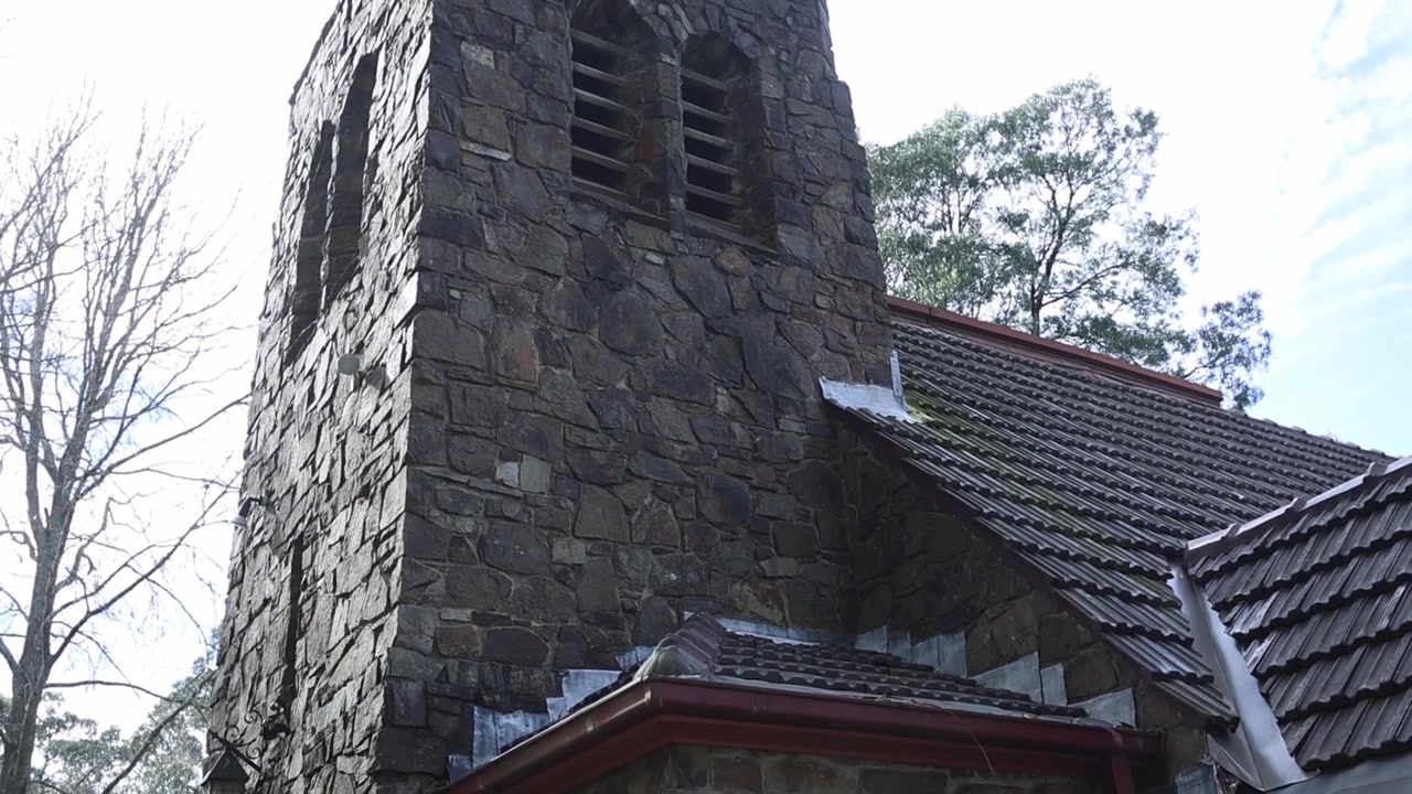 Lavvinkelbillede af en gammel stenkirke i Olinda, Melbourne, med detaljeret murværk, et vejrbidt klokketårn, løvtræer og overskyet dagslys for en fredfyldt, tidløs stemning.