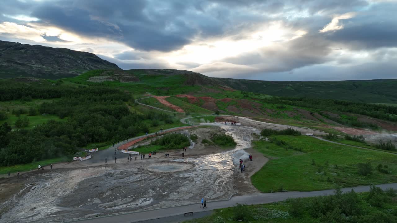 아이슬란드의 지열지역인 게이시르 (geysir) 를 걸어다니는 방문객들의 드론 풍경, 초록색 언덕에 둘러싸여 있고 해가 지는 동안 극적인 하늘.