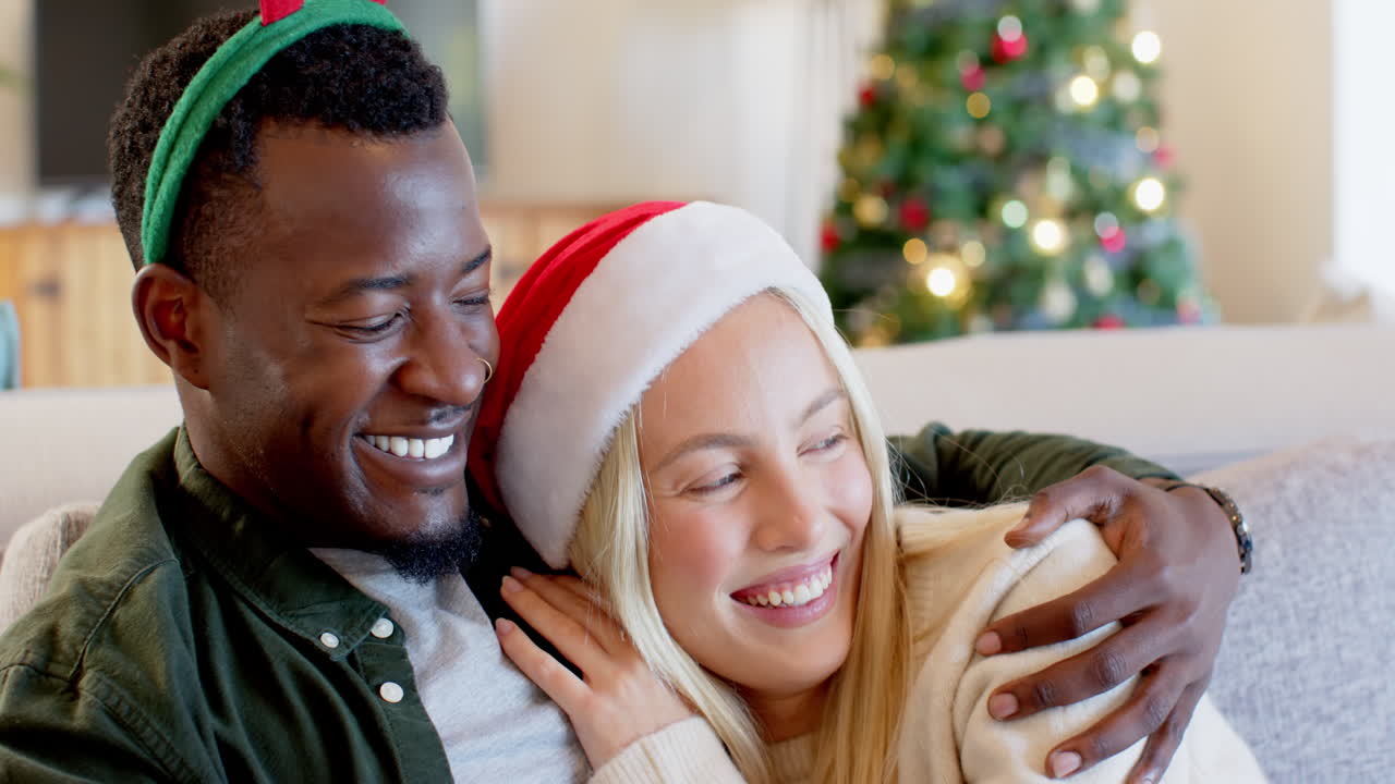 Smiling diverse couple embracing on couch with Christmas tree in background