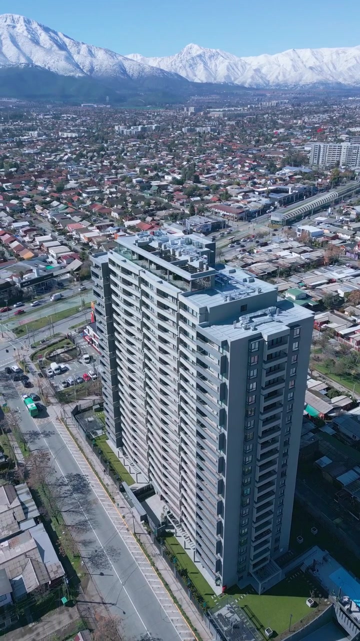 Volo circolare intorno a un'alta torre residenziale con la città che si estende e la catena montuosa delle Ande innevate sullo sfondo. Santiago, Cile.