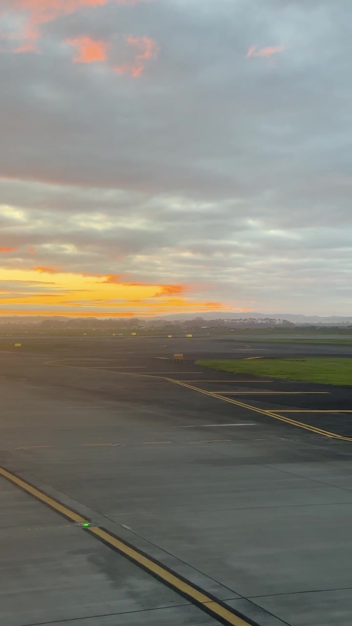 Pesawat melaju di landasan pacu Auckland saat matahari terbenam. Langit yang cerah, gerakan kamera yang halus, suasana yang tenang.