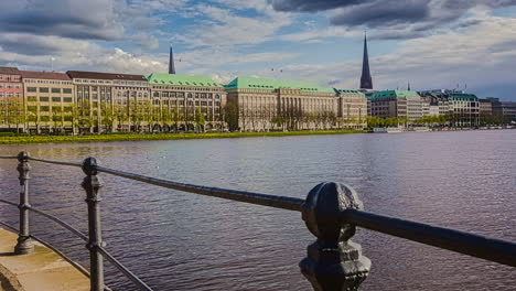 Tiro-De-Hiperlapso-Del-Lago-Alster-De-Hamburgo-Con-Fuente-Y-Hermosos-Edificios-Históricos-En-La-Costa-Durante-El-Día-Nublado---Hamburgo,-Alemania