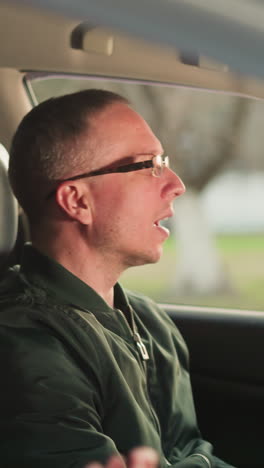 una joven con una gorra rosada y una chaqueta está sentada en el asiento delantero del pasajero de un coche, mientras que un hombre, con gafas y unachaqueta verde, está en el asiento del conductor, cantando con entusiasmo