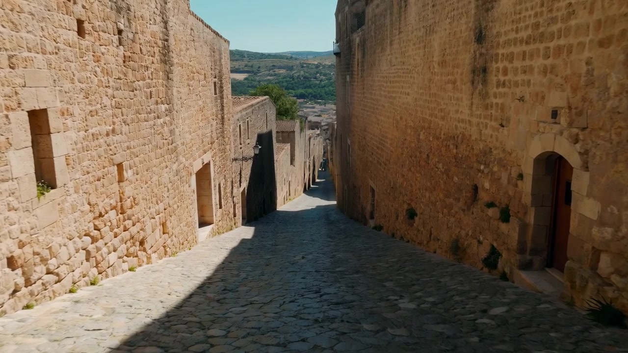 Vista dall'alto lungo una stretta via acciottolata fiancheggiata da imponenti facciate in arenaria stagionata, con un'ombra diagonale che si muove in primo piano mentre la valle distante e le cime degli alberi rimangono visibili sotto un cielo limpido.