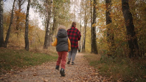 Una-Mujer-Joven-Y-Un-Niño-Pequeño-Corren-En-El-Bosque-Otoñal-Sobre-Un-Sendero.-El-Hijo-Sigue-A-La-Madre-Para-Ponerse-Al-Día-Con-La-Naturaleza-Durante-El-Fin-De-Semana.-Vista-Trasera-De-Una-Familia-Feliz.