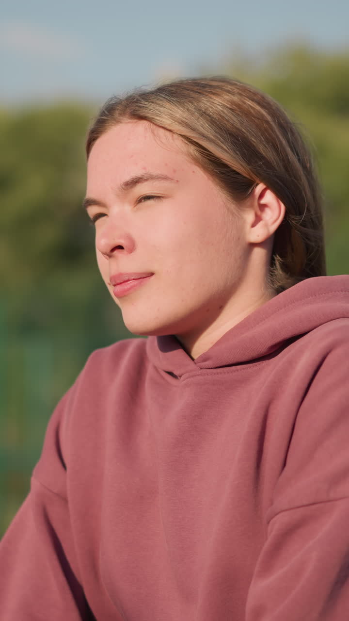 피트니스 운동에 참여하는 운동선수, 앞으로 무을 고 머리카락을 뒤로 고, 야외 스포츠 환경에서 나무와 불분명한 인물이 있는 흐릿한 배경으로 아침 드릴을 즐기고 있습니다.