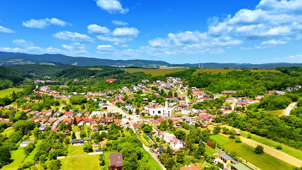 Tranquil village in greenery. Aerial view of a picturesque village with houses nestled amid lush hills and vibrant fields under a bright blue sky
