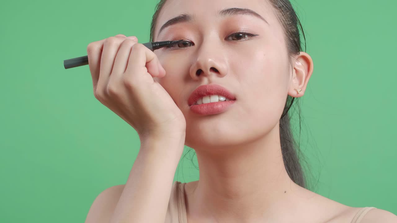Close Up Of Asian Woman Applying Eyeliner Doing Perfect Eye Arrows And Smiling While Standing On Green Screen Background In The Studio