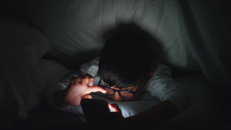 Primo piano di un ragazzo nella sua camera da letto a casa che usa il telefono cellulare per inviare messaggi sotto le coperte o il piumone di notte 8