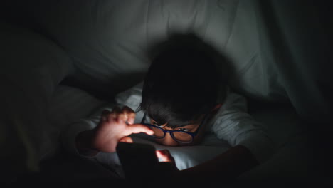 집에서 침실에 있는 어린 소년의 클로즈업은 밤 8시 개나 침대 에서 드폰으로 문자 메시지를 보내고 있다.