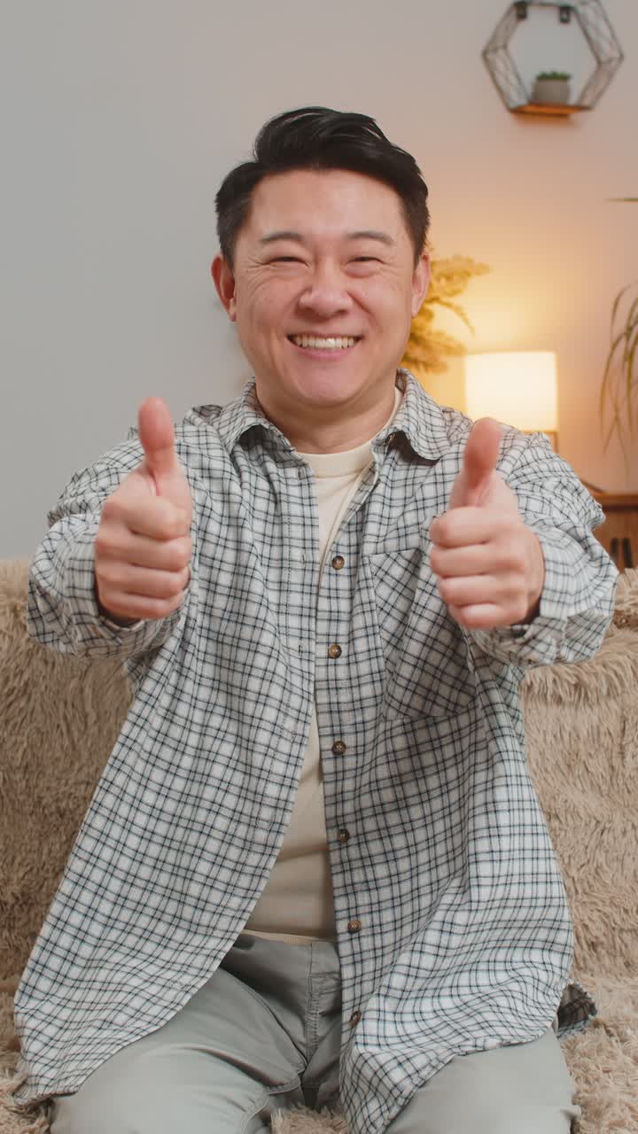 Portrait of excited smiling chinese young man showing double thumbs up sitting on sofa couch at home