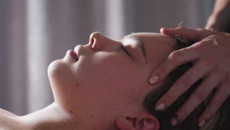close up of woman with light skin and closed eyes lying down while receiving gentle head massage, therapist's hand softly pressing on forehead in calm environment with soft natural lighting