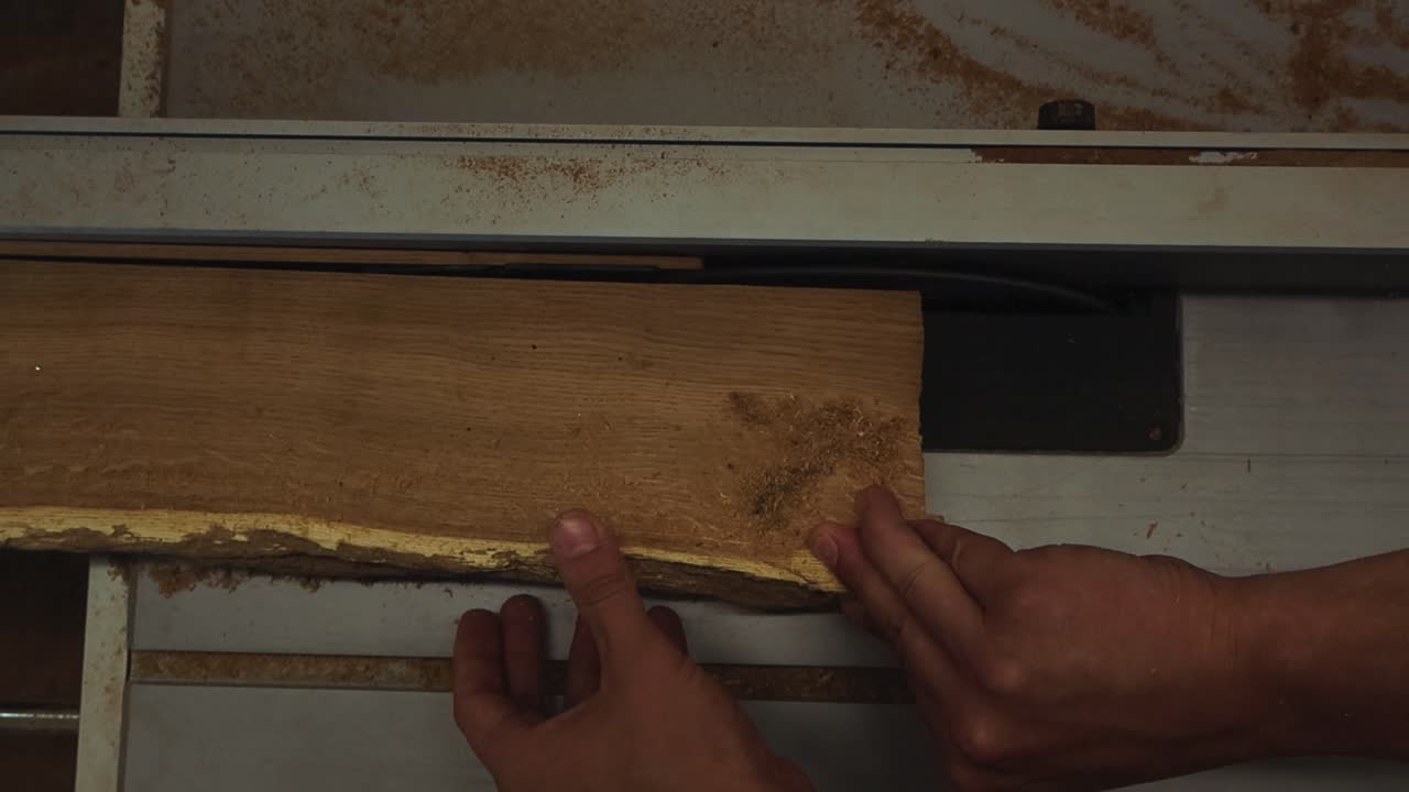 Top down view of a diverse person hands pushing a oak slab or timber through a table saw while the blade is cutting thing slices off it in slow motion. Saw dust and wood chips fly around. No gloves