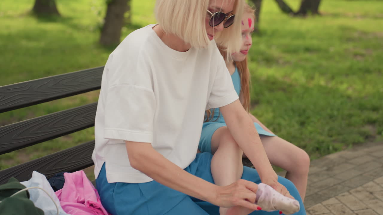 mother seated beside daughter on bench in park putting sock on child foot, casual summer clothes, green background, caring moment between parent and child sharing smile and connection