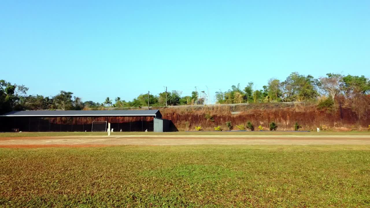 Wide drone shot of a quiet cricket ground and training nets under a clear blue sky, featuring a lush green field and a rustic red earth backdrop