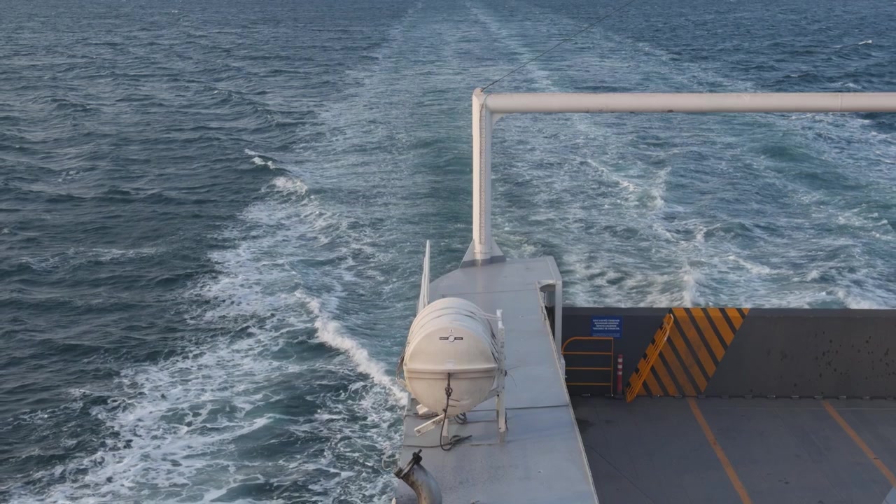Statische Weitwinkelaufnahme von einer fahrenden Fähre, die ein Frachtschiff im Marmarameer zeigt, mit der Küstenlinie Istanbuls im Hintergrund während der goldenen Stunde.