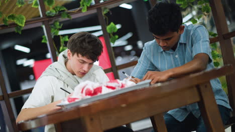 moving view capturing youngsters paying full attention on work with tray of chips on table while hands sketch on paper collaborative study session in cosy indoor setting with snack break time fun vibe