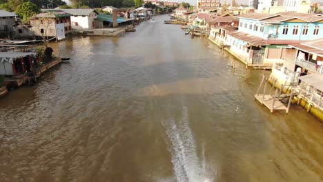 Boat-driving-on-river-in-Nyaung-Shwe-Myanmar-at-Inle-Lake,-traditional-buildings-in-background