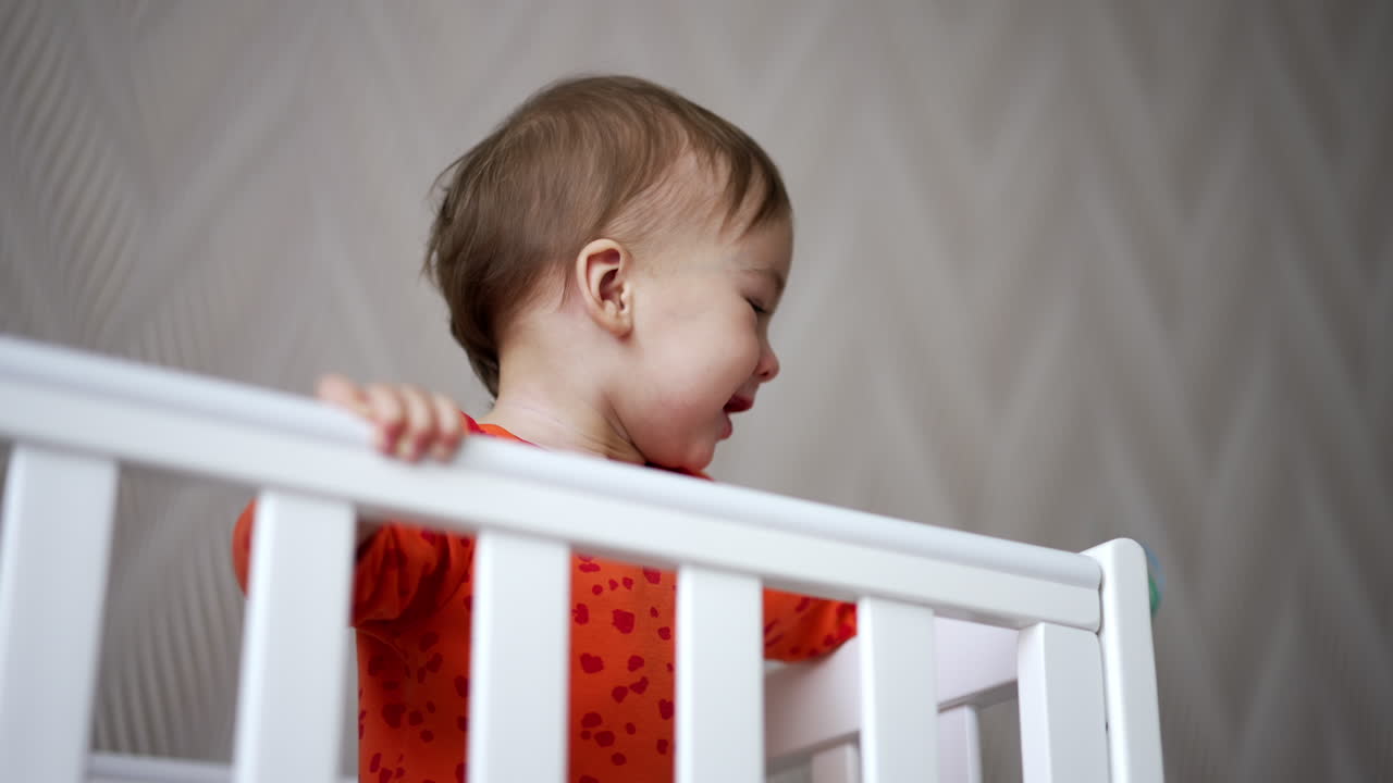 Active Caucasian kid stands up in his cot holding by the handrail. Smiling baby waving hand with pacifier. Low angle view.