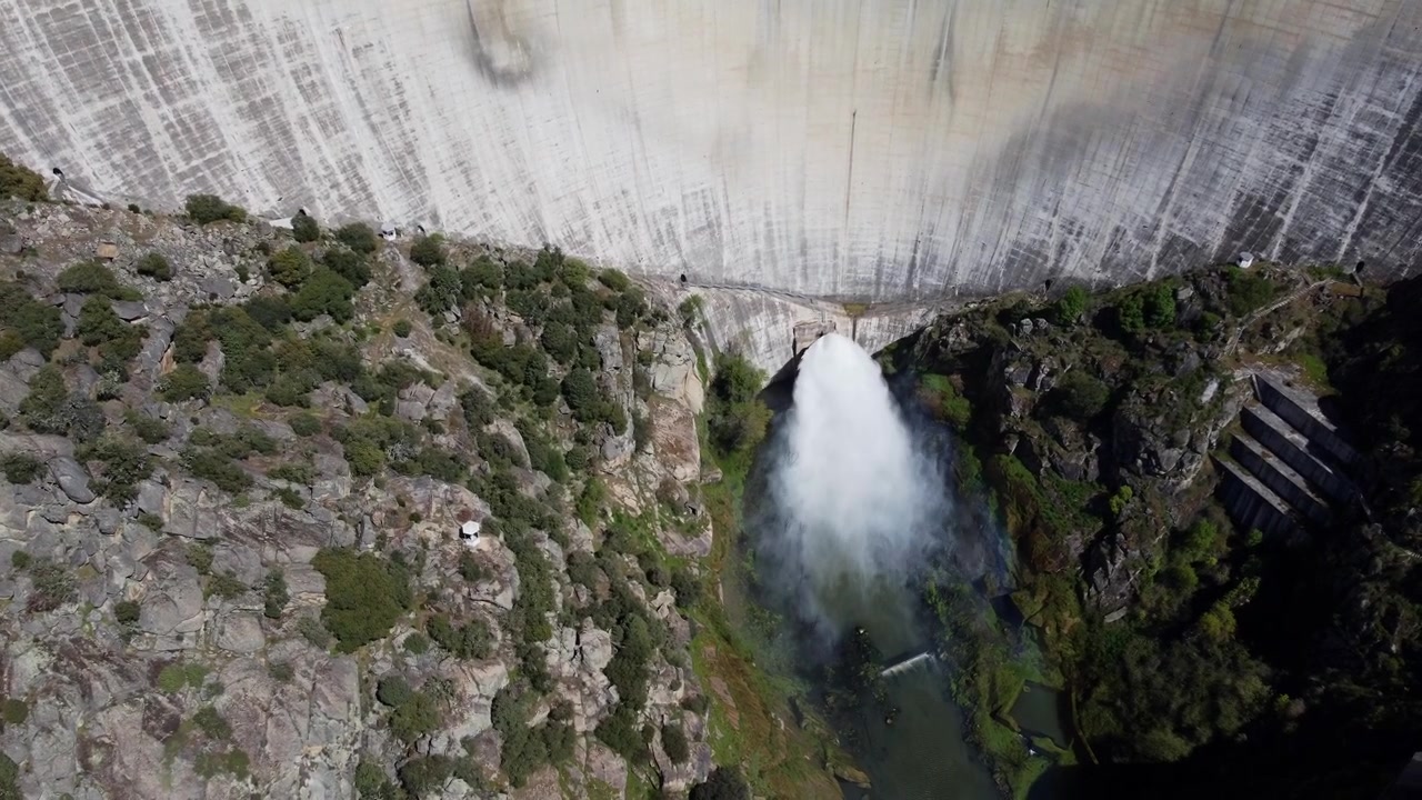 4K aerial drone video descending towards the monumental Almendra Dam in Salamanca, Spain. Focused on a powerful gush of water, released under pressure, shooting from the concrete spillway into the riv