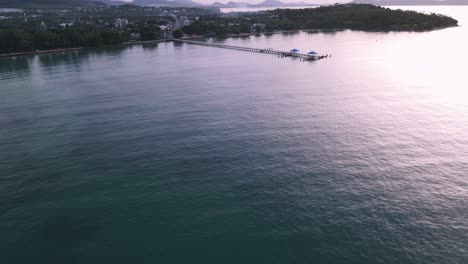 Panorámica-Aérea-De-Drones-Sobre-Aguas-Azules-Tropicales-Hasta-El-Muelle-Durante-El-Amanecer-En-La-Ladera-De-Una-Montaña-Verde