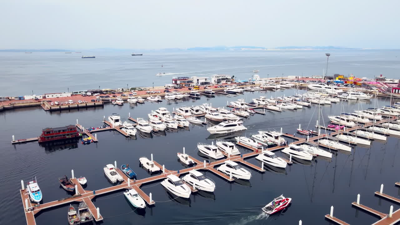 Luftfoto av en marina i skumringen med flere luksusbåter, yachter og katamaraner fortøyd til brygger i marinaen, i kontrast til byens silhuett og turkise vann i bakgrunnen
