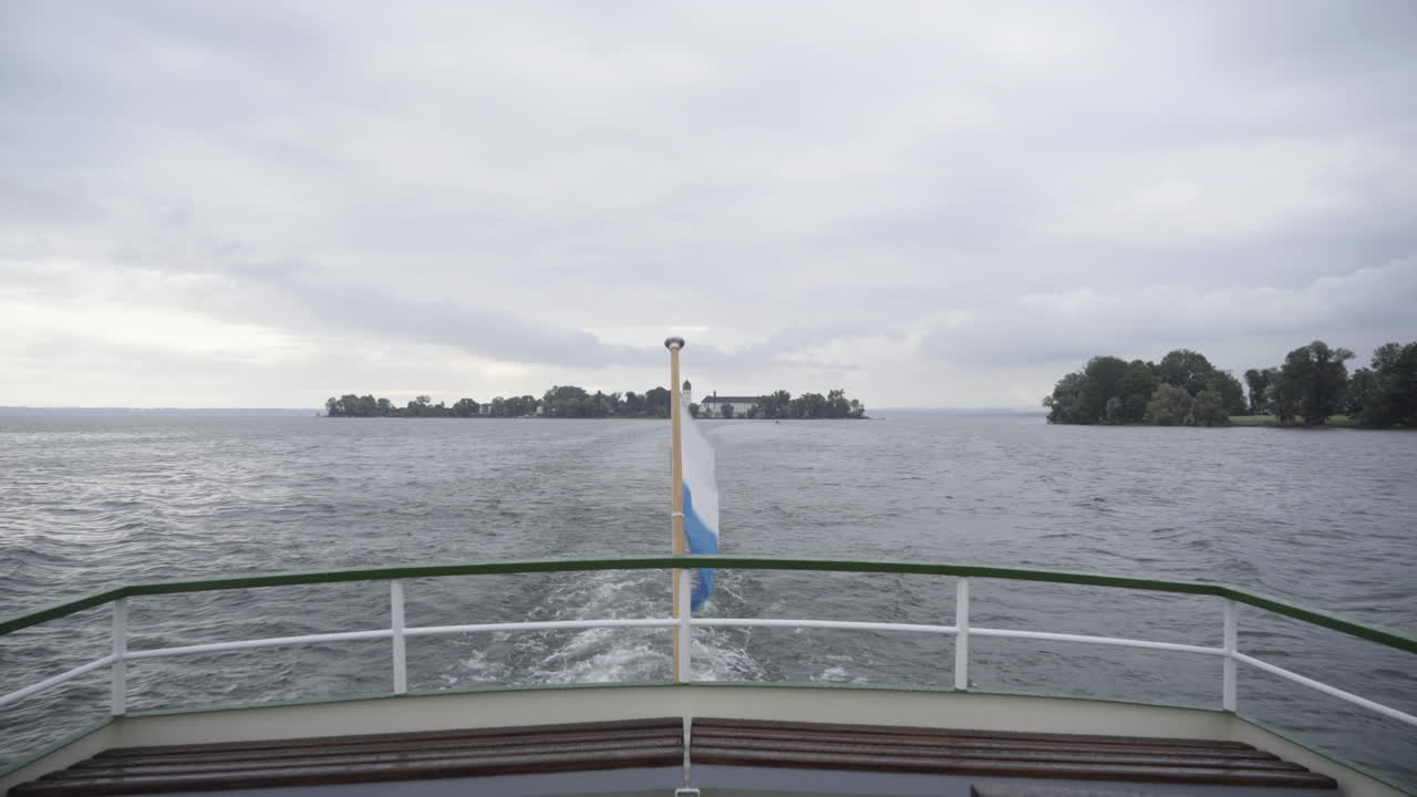 Eine ruhige Gimbal-Aufnahme vom Heck eines Bootes, mit einer Flagge im Vordergrund und einer Insel im Hintergrund auf dem Chiemsee.