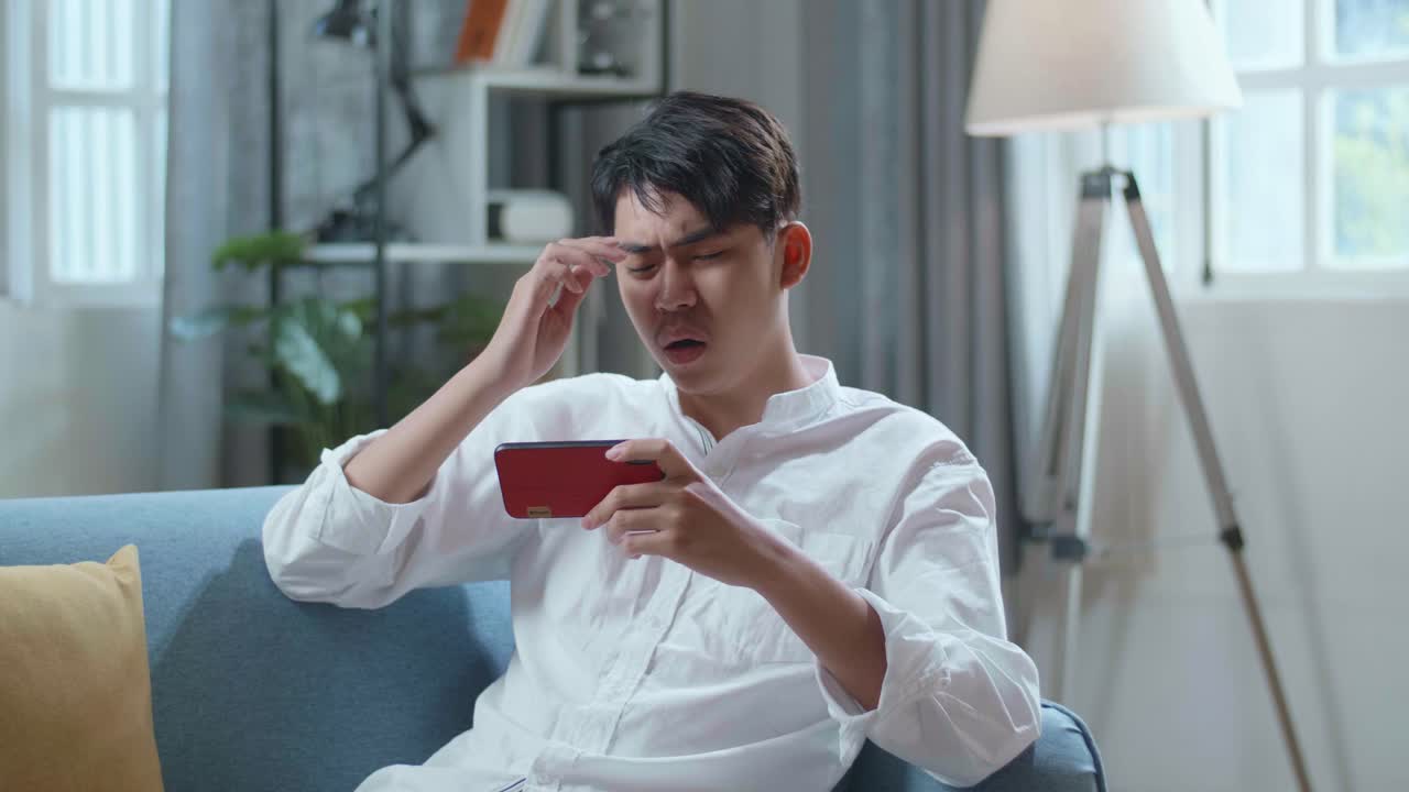 Asian Man Being Upset Losing Game On Smartphone While Lying On Sofa In The Living Room