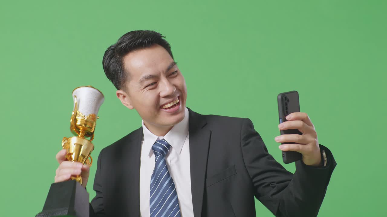 슈트와 타이를 입은 아시아 사업가와 금메달과 금 트로피를 보여주는 가까운 사진 스마트폰에서 녹색 화면 배경으로 스튜디오에서 비디오 통화