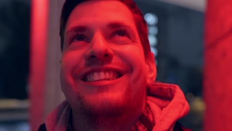 joven feliz mirando hacia arriba y sonriendo en luz de neón roja, primer plano en la cara en vista orbital de mano