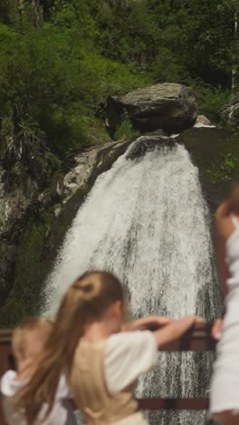 high waterfall with white sprays and happy family on bridge in wild park. woman with little children enjoy fresh cascade on mountain river in forest